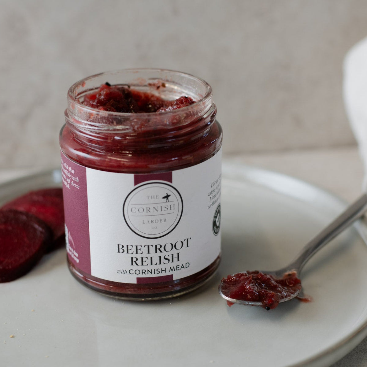 A jar of Beetroot relish sits on a light gray ceramic plate, accompanied by sliced beetroot, a spoonful of the relish, and a white cloth in the background.