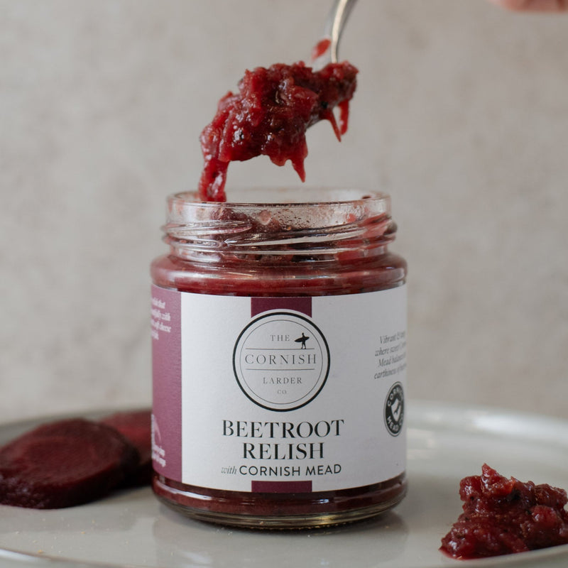 A hand with pink nail polish holds a spoonful of dark red beetroot relish above an open glass jar labeled 'The Cornish Larder Beetroot Relish with Cornish Mead'.