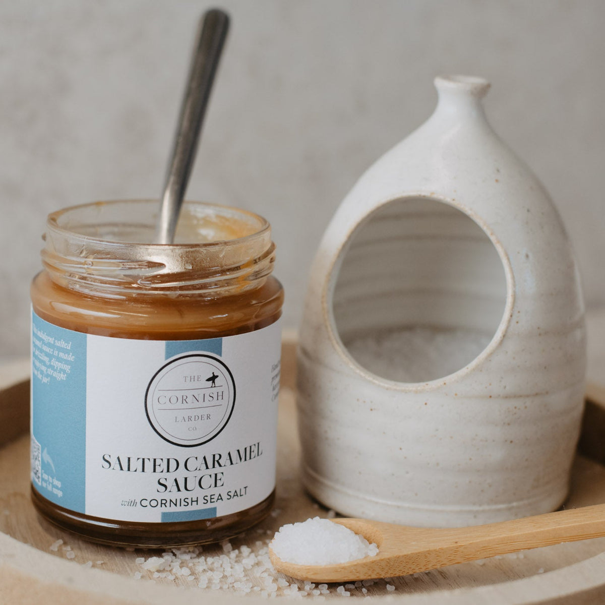 A jar of Salted Caramel Sauce sits on a round wooden tray next to a ceramic salt dish and a wooden spoon with Cornish Sea Salt, against a neutral textured background.