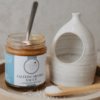 A jar of Salted Caramel Sauce sits on a round wooden tray next to a ceramic salt dish and a wooden spoon with Cornish Sea Salt, against a neutral textured background.