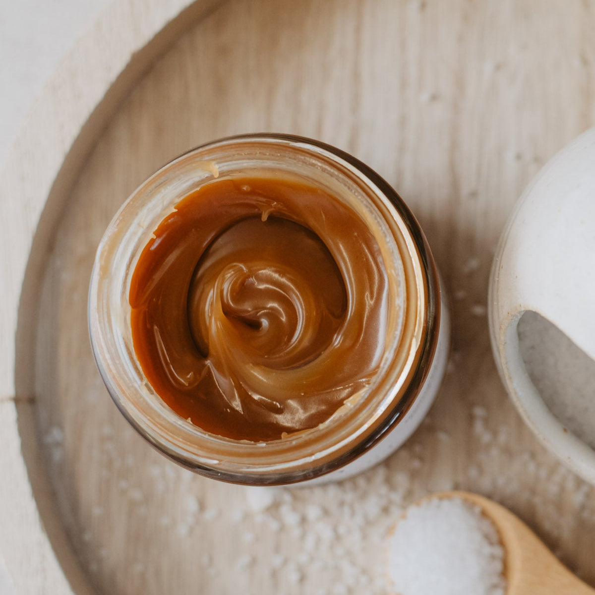 A top-down view of a glass jar filled with caramel sauce, placed on a light wood tray with scattered sea salt and a wooden spoon holding salt nearby.