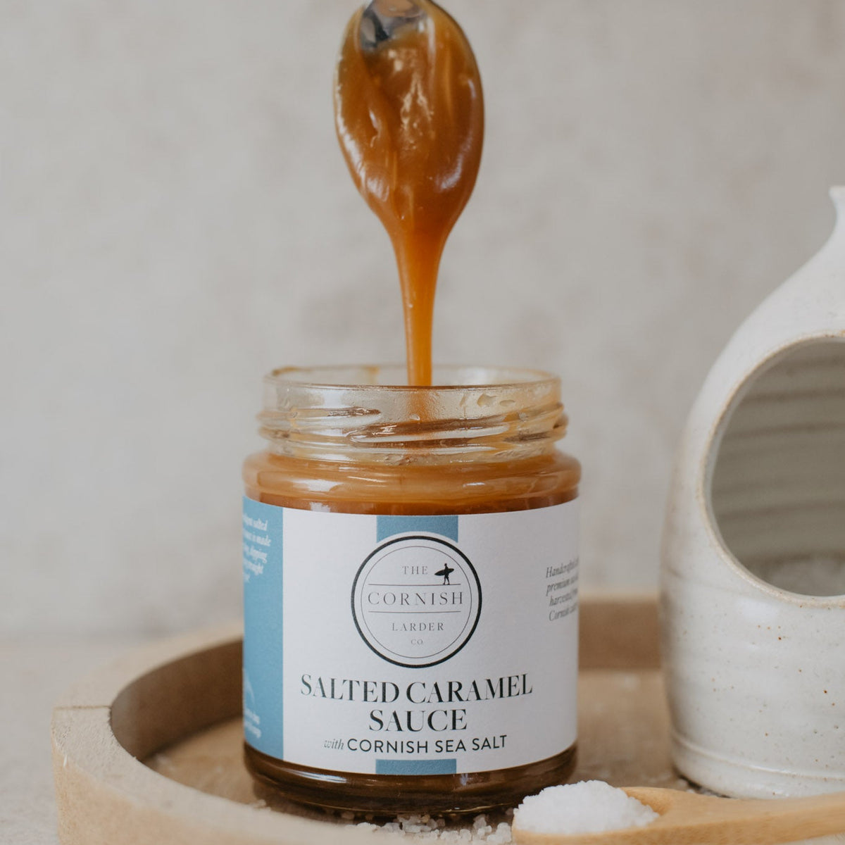 A glass jar of salted caramel sauce sits on a wooden tray, with caramel being drizzled from a metal spoon into the jar by a hand with red nail polish. A white ceramic dish is partially visible on the right, and the background is a soft, neutral textured wall.