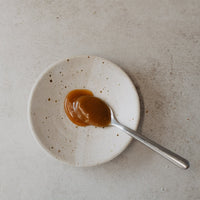 A spoonful of glossy caramel sauce resting on a small speckled white ceramic plate, set against a neutral textured light gray background.