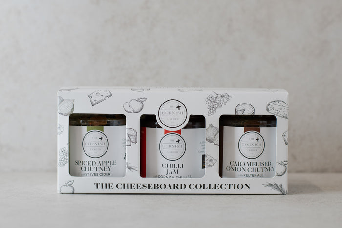 A boxed three-pack of gourmet spreads labeled 'The Cheeseboard Collection,' showing three jars through a white printed package: Spiced Apple Chutney, Chilli Jam, and Caramelised Onion Chutney.