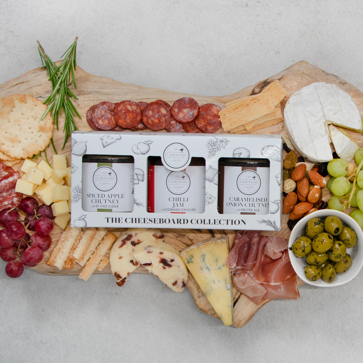 An overhead flat-lay of a charcuterie board styled with cheeses, crackers, salami, grapes, olives, nuts, and a boxed set of three cheese boards chutneys from The Cheese Larder, set on a light gray surface.