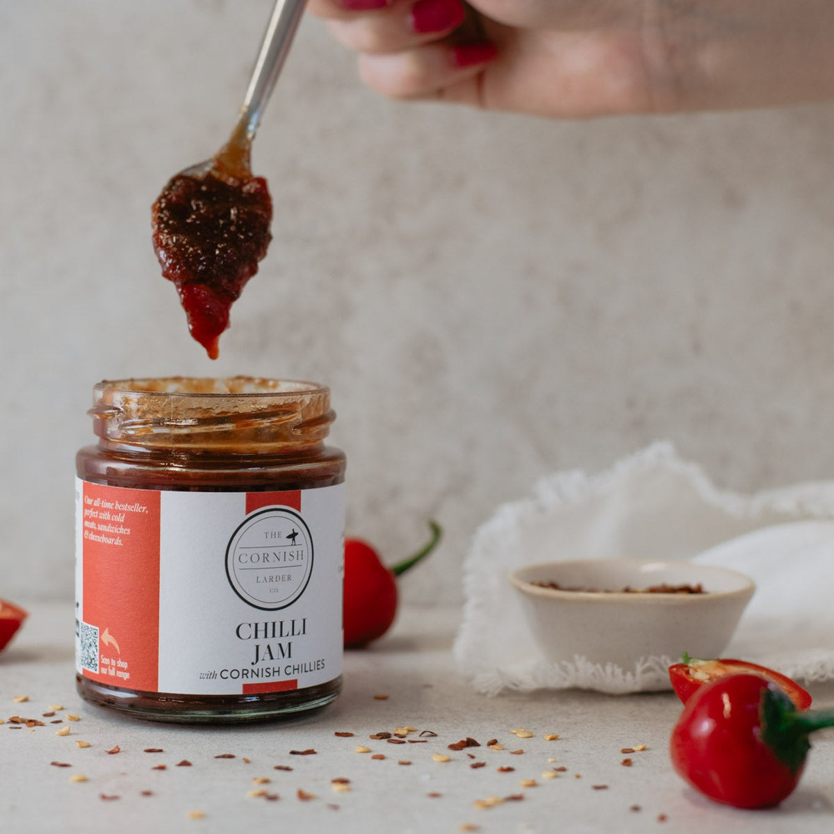 A jar of 'Chilli Jam' by The Cornish Larder is shown with a spoon lifting a dollop of the chilli jam above it. The scene is styled with fresh red chili peppers and scattered chilli flakes on a neutral, textured background.