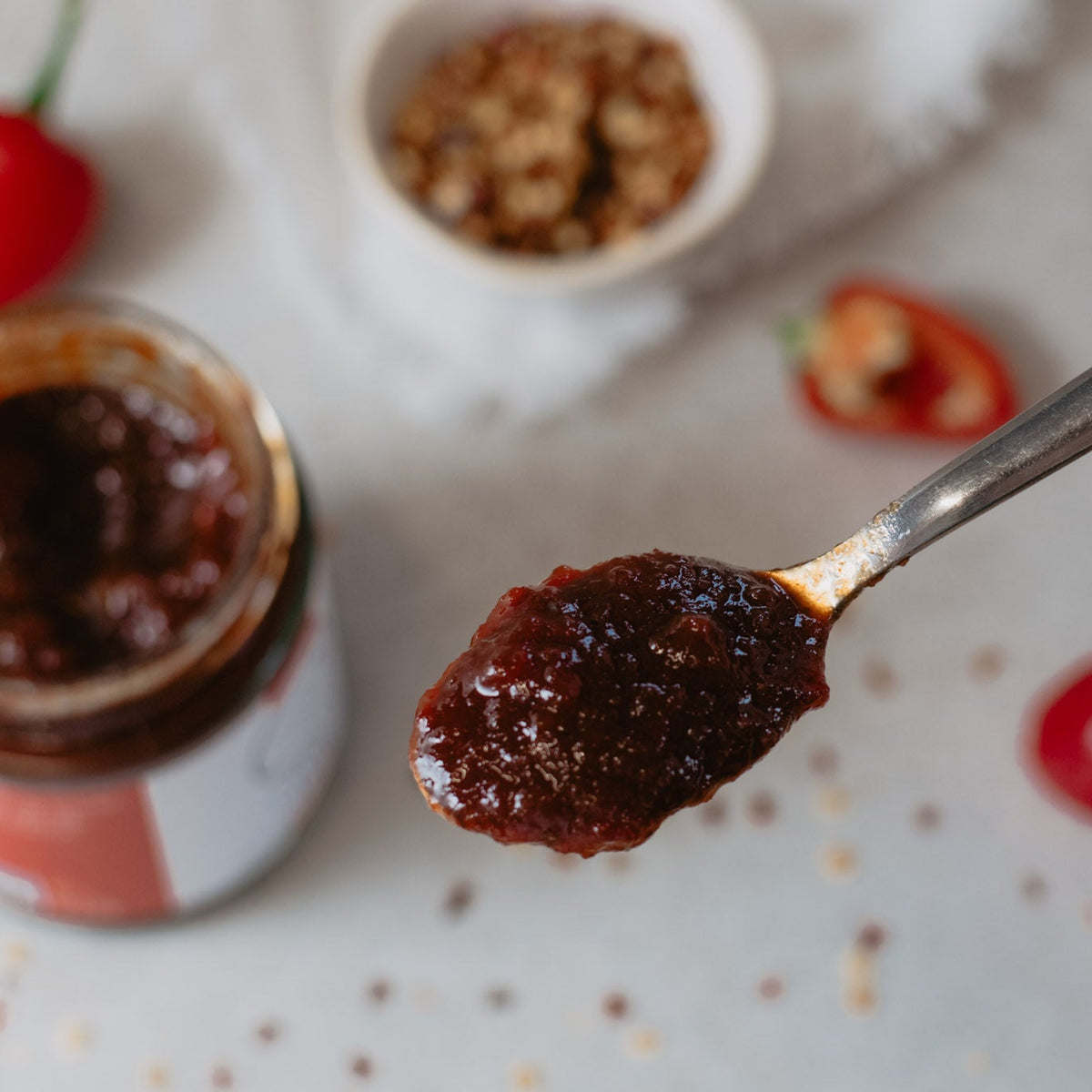 A close-up of a hand holding a spoon filled with a thick, dark red chili jam in front of an open jar of the same spread, with small red chili peppers and a bowl of seasoning scattered on a light surface.