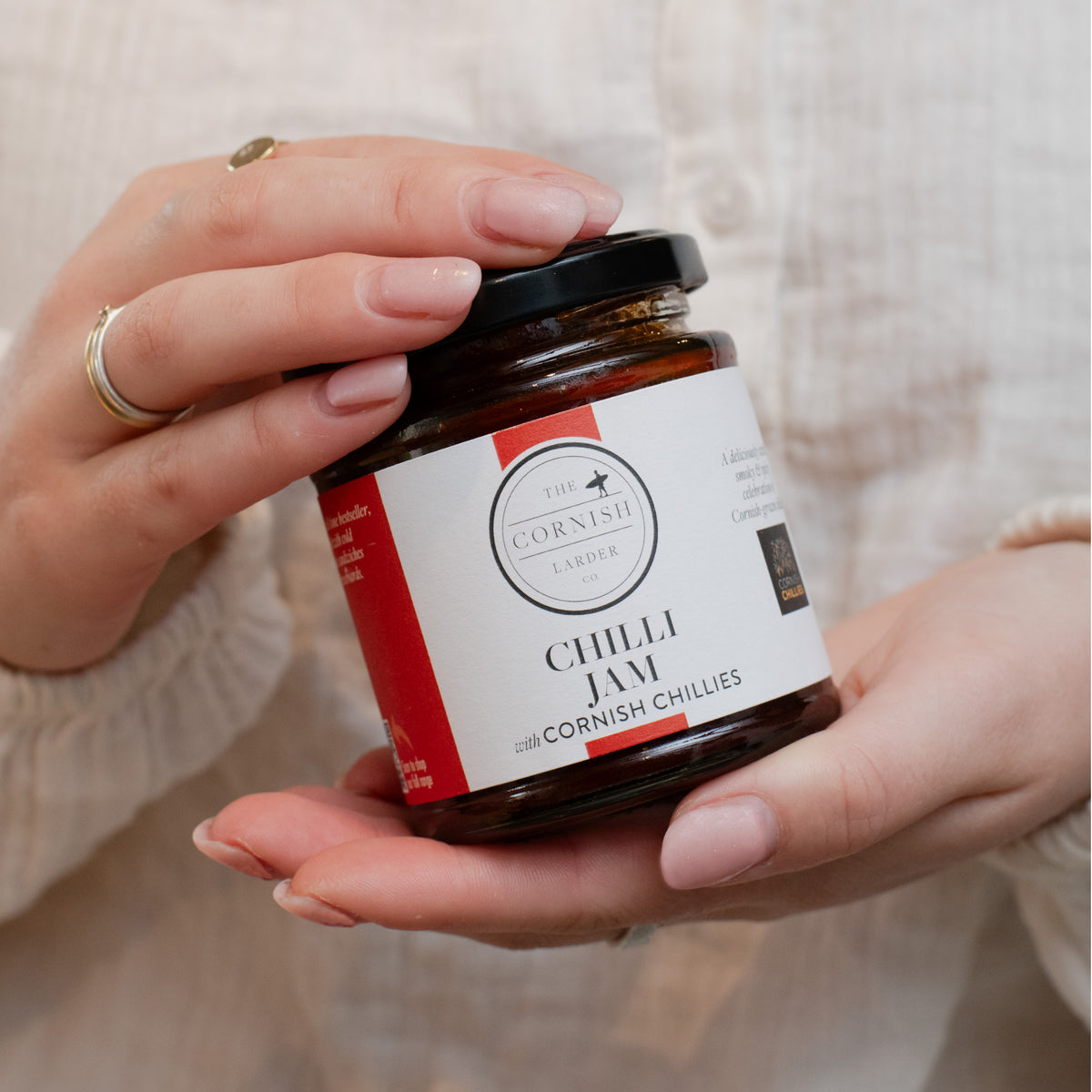 A person holds a jar of Chilli Jam with Cornish Chilies at an angle. The jar has a black lid and a white label with red accents, featuring the brand 'The Cornish Larder'.