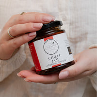 A person holds a jar of Chilli Jam with Cornish Chilies at an angle. The jar has a black lid and a white label with red accents, featuring the brand 'The Cornish Larder'.
