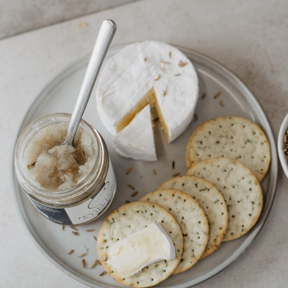 An overhead flat-lay of a cheese and cracker spread on a light stone surface. A round white cheese with a wedge cut out sits on a gray platter beside a jar of fennel, lemon and tarragon relish with a spoon, several herb-cracker rounds, and a small white bowl of fennel seeds.