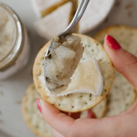 A close-up lifestyle shot of a hand with pink nail polish spreading a fennel, lemon and tarragon relish on a round cracker. 