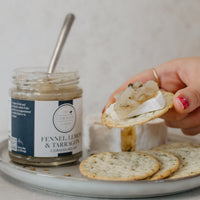A hand holds a round cracker topped with brie and a dollop of fennel, lemon and tarragon relish with the jar in the background on a plate with more crackers.