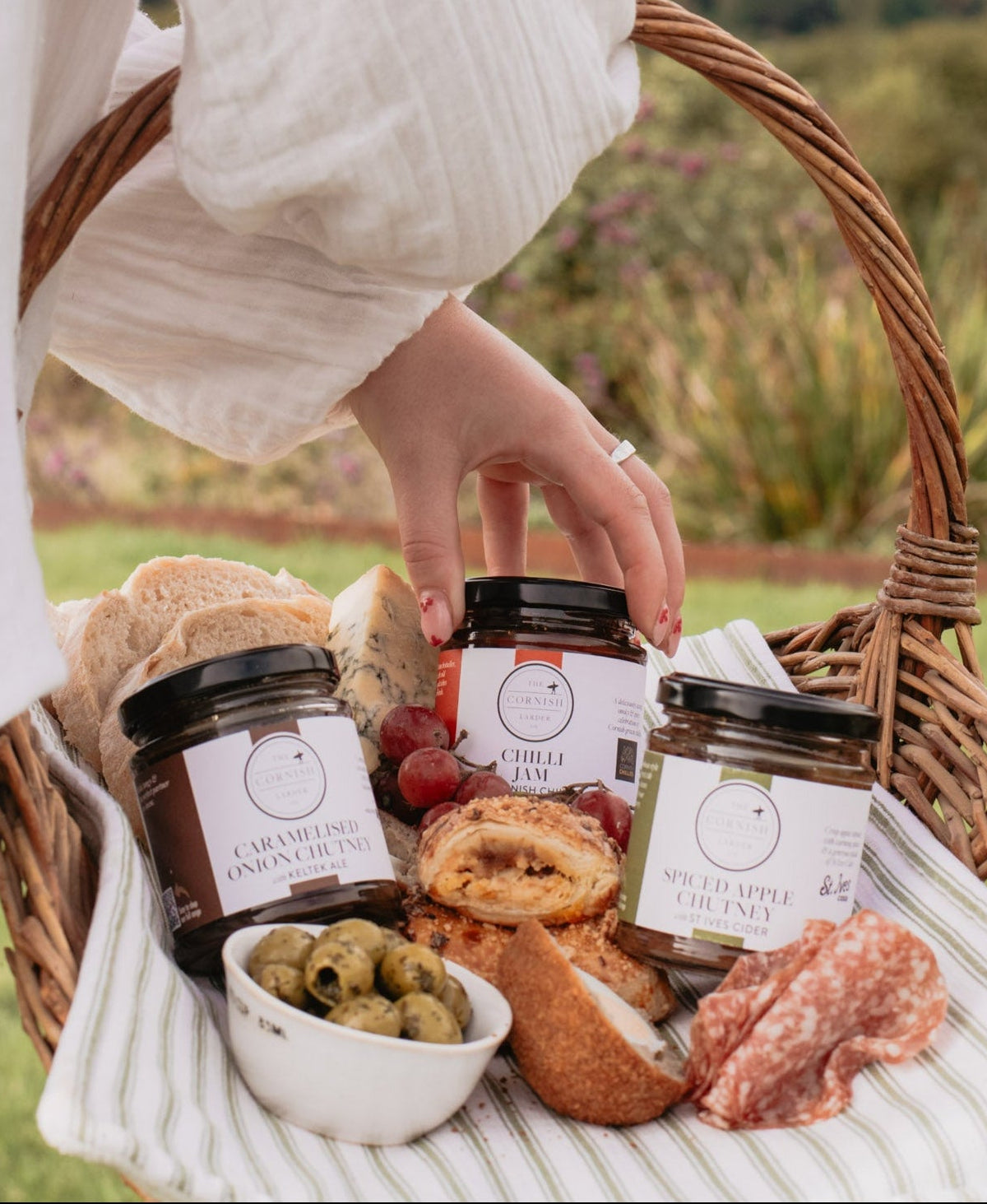 A wicker picnic basket filled with assorted gourmet foods, including jars of spreads, bread, cheese, olives, and salami, held by a person in a white blouse against a blurred outdoor landscape background.