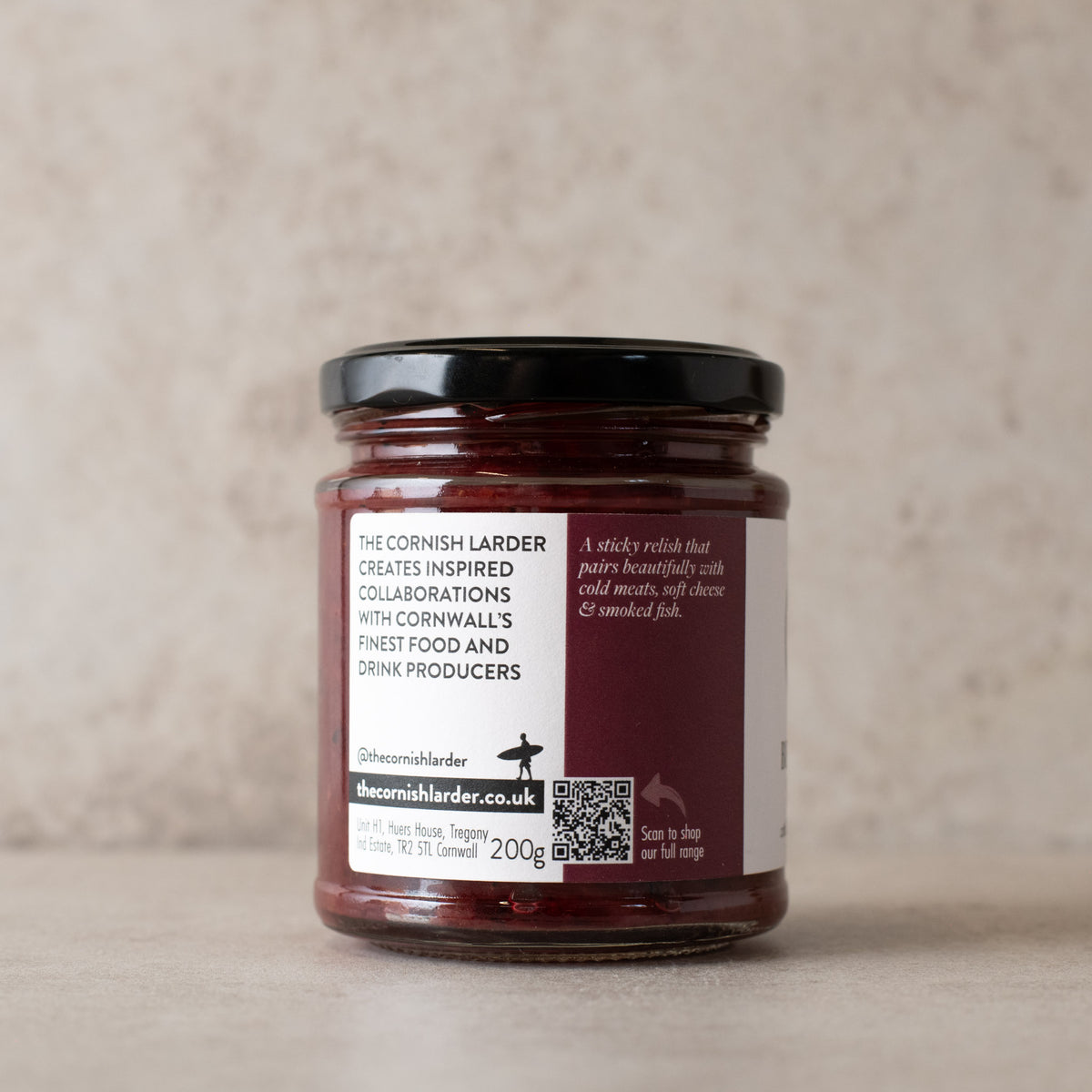 A 200g glass jar of The Cornish Larder beetroot relish, featuring a black lid and a white label with dark red text, set against a neutral textured beige background.