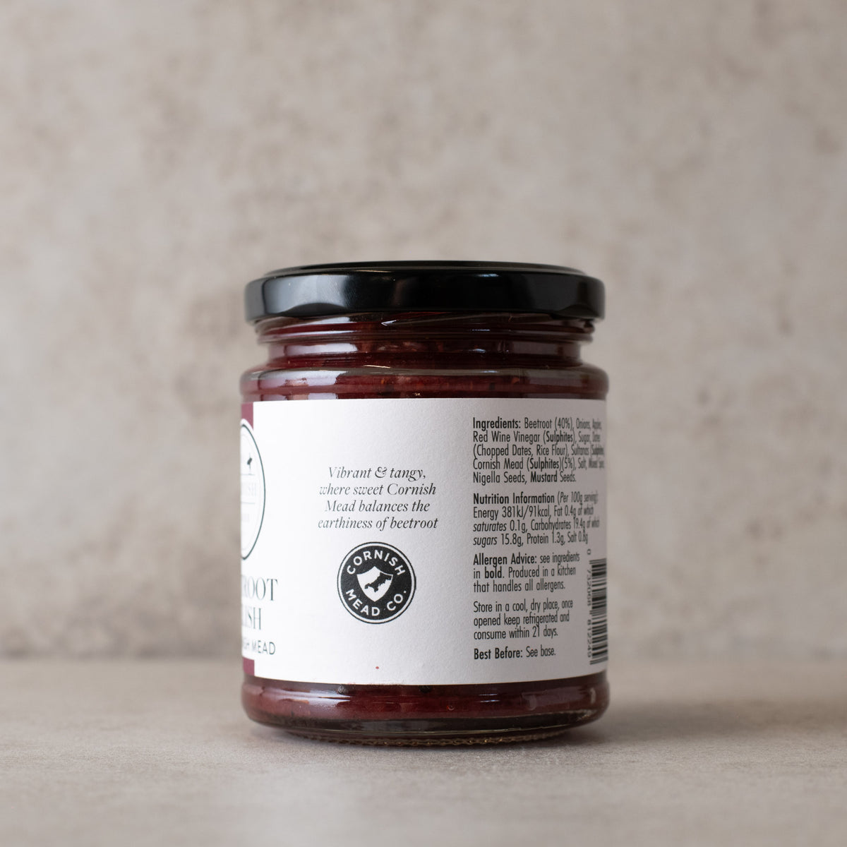 A glass jar of beetroot relish with a black lid and a white label, showing one side of the label with ingredients and nutrition details, set against a neutral beige background.