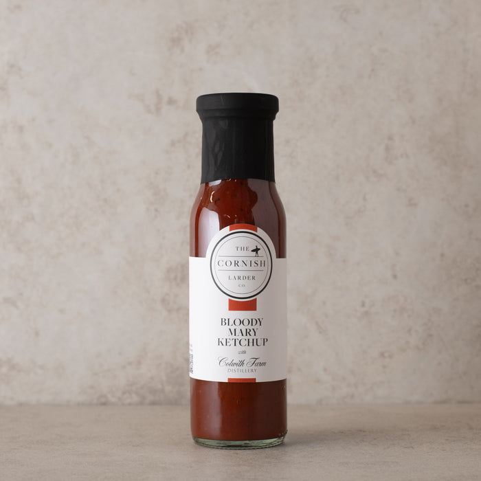 A bottle of Bloody Mary Ketchup from The Cornish Larder, featuring a white and red label with black text, a black cap, and reddish-brown sauce inside, set against a textured beige background.