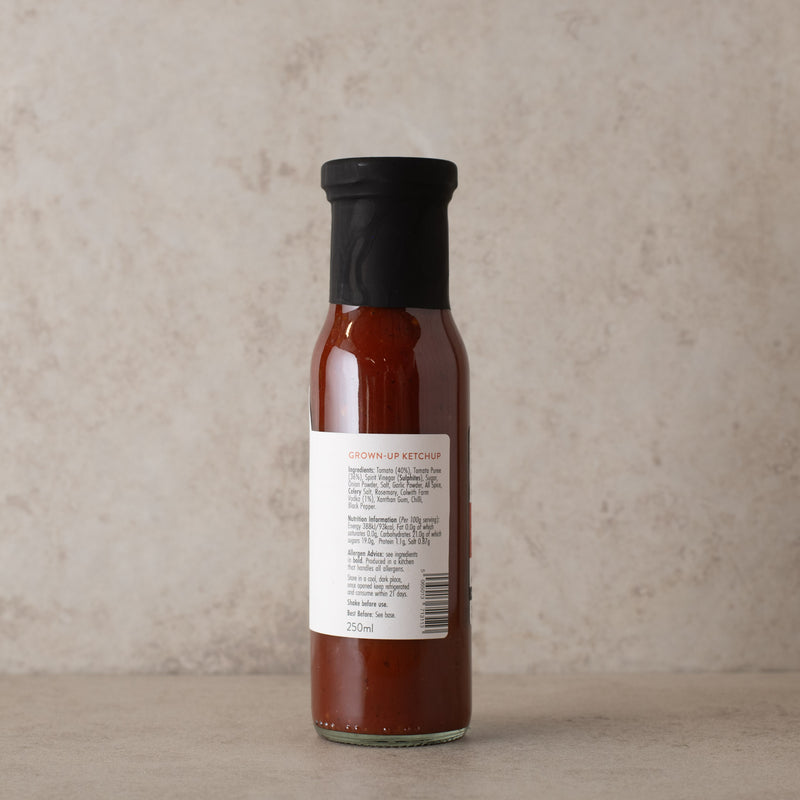 Side shot of Bloody Mary Ketchup bottle with a black cap and a white label, standing on a beige surface against a textured beige background.