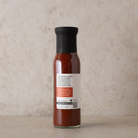 A side shot of a bottle of Cornish Larder ketchup with a black cap and white label, set against a textured beige background.