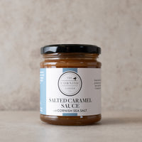 A  jar of The Cornish Larder Salted Caramel Sauce with a black lid and white label, set against a neutral textured beige background.