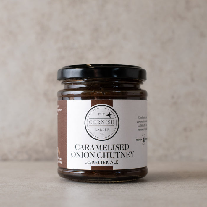 A glass jar of 'The Cornish Larder' Caramelised Onion Chutney with a black lid and a white label, set against a neutral textured beige background.