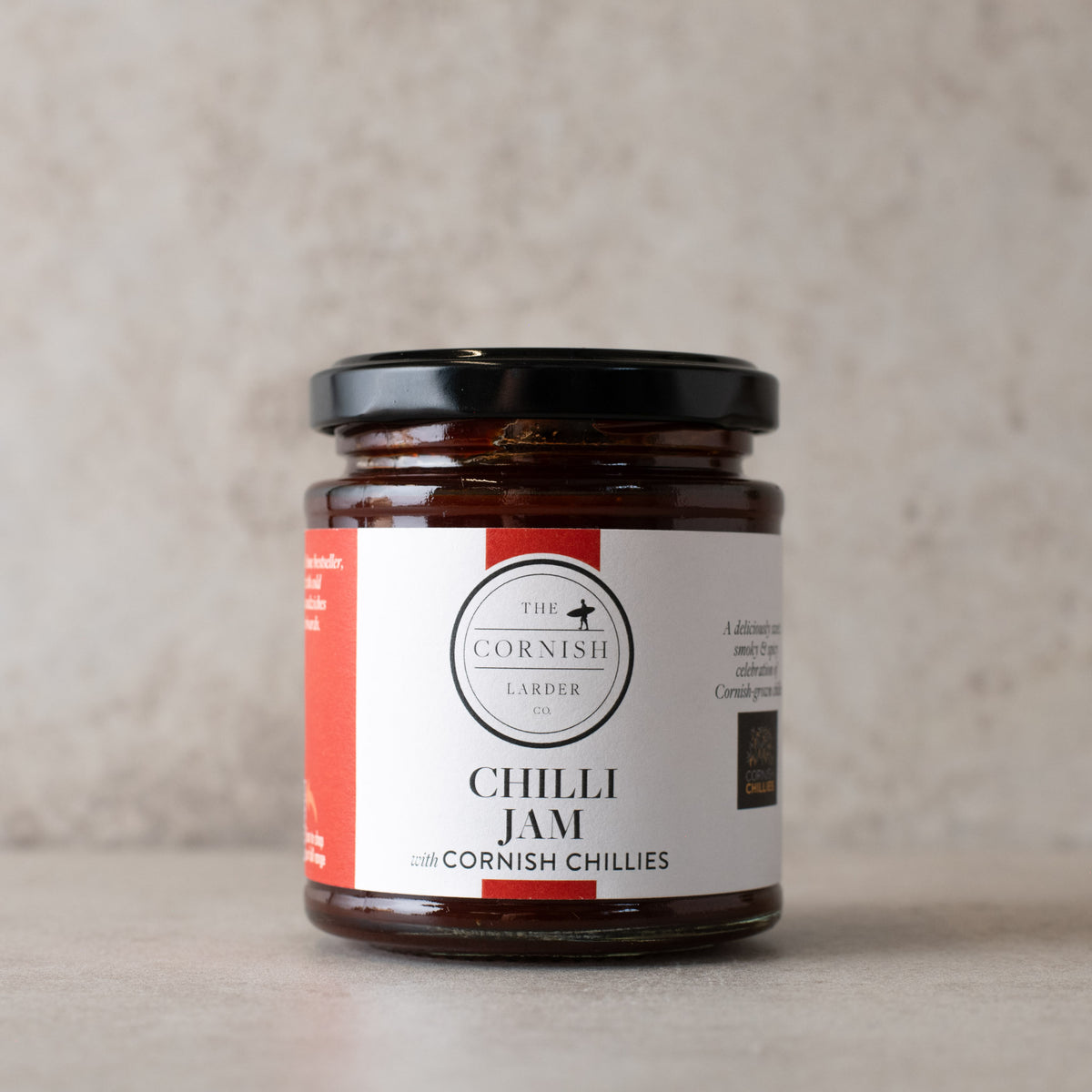 A glass jar of Cornish Larder Chilli Jam with a black lid and a white label with red accents, set against a neutral textured beige background.