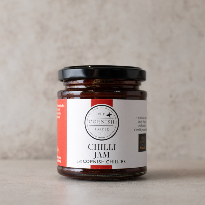 A glass jar of Cornish Larder Chilli Jam with a black lid and a white label with red accents, set against a neutral textured beige background.
