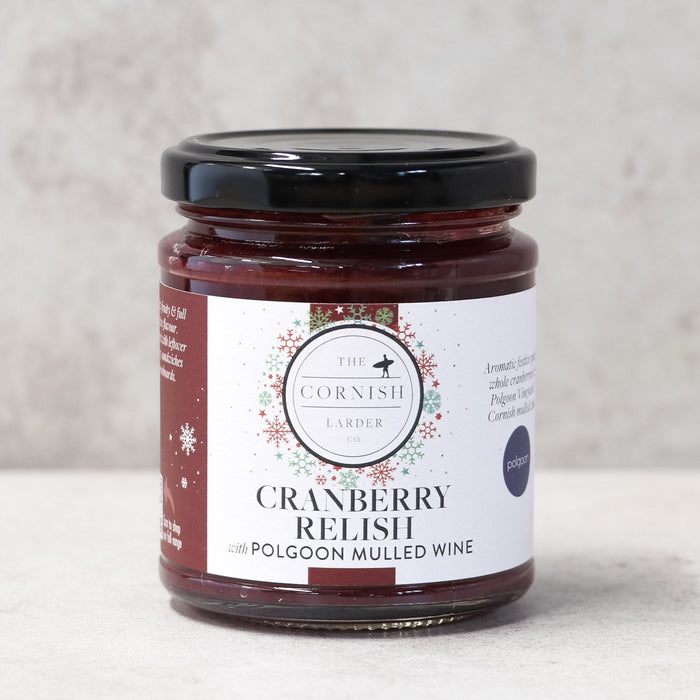 A glass jar of The Cornish Larder Cranberry Relish with a black lid, set against a neutral, light beige textured background.