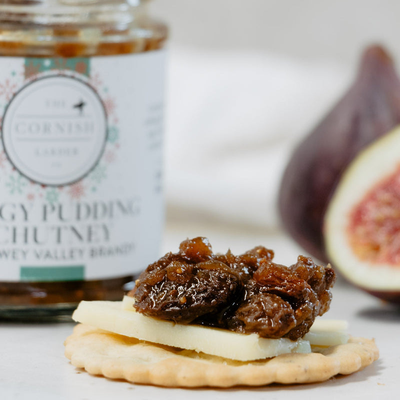 A jar of Cornish Larder Figgy Pudding Chutney with a spoon, shown next to a cracker topped with cheese and figs on a light background.