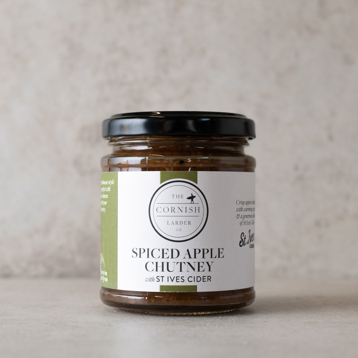 A glass jar of The Cornish Larder Spiced Apple Chutney with a black lid and white label, set against a neutral textured beige background.