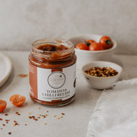 A glass jar of The Cornish Larders Tomato & Chilli Relish sits on a light stone surface, surrounded by halved cherry tomatoes, bowls of seasoning, and a white cloth in a softly lit kitchen setting.