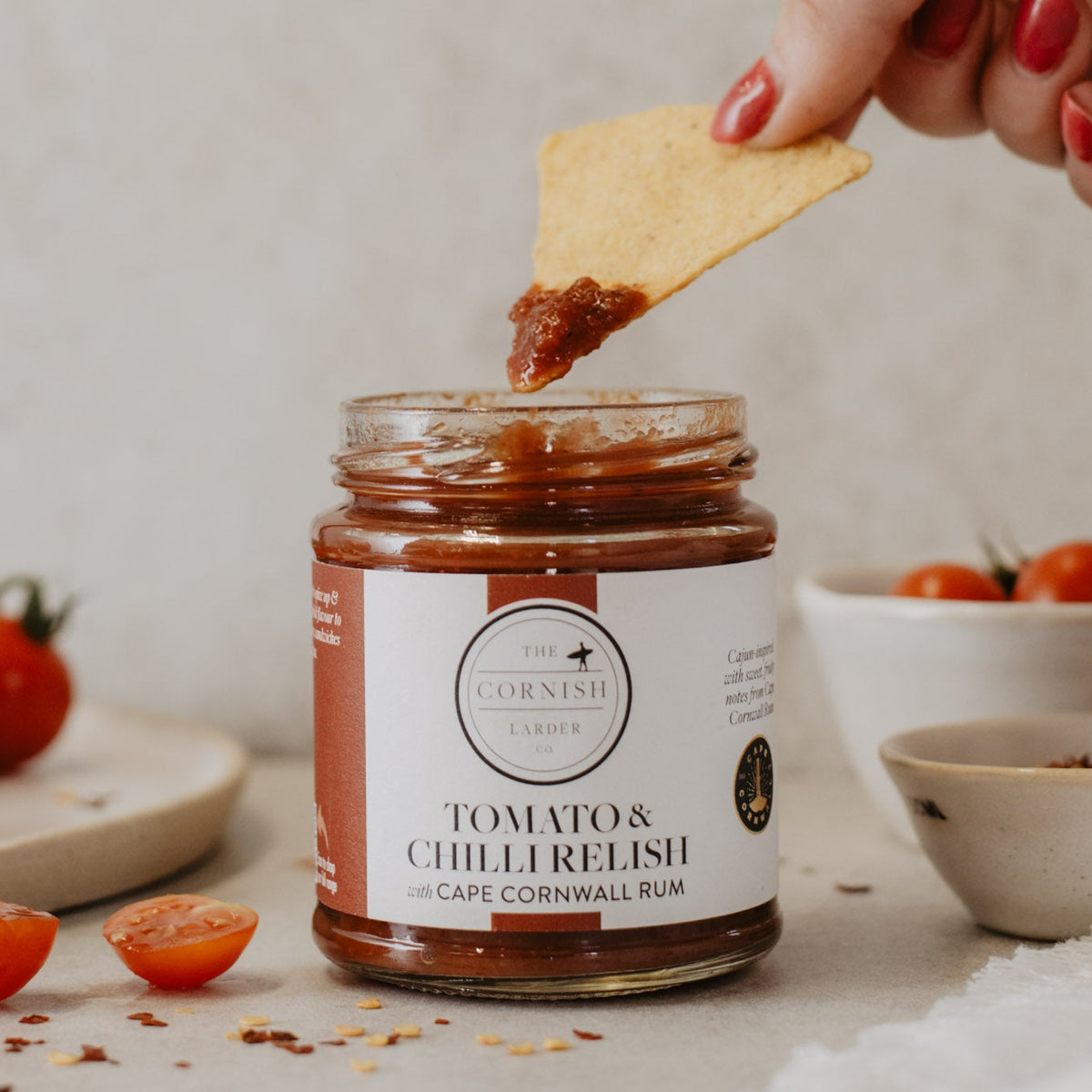 A glass jar of Tomato & Chilli Relish by The Cornish Larder, with a hand dipping a tortilla chip in. Fresh cherry tomatoes are scattered on a light stone surface in the background.