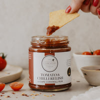 A glass jar of Tomato & Chilli Relish by The Cornish Larder, with a hand dipping a tortilla chip in. Fresh cherry tomatoes are scattered on a light stone surface in the background.