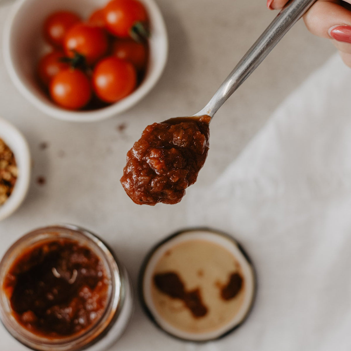 A close-up shot of a hand holding a spoon filled with a chunky red tomato relish with bowls of cherry tomatoes and spices in the background.