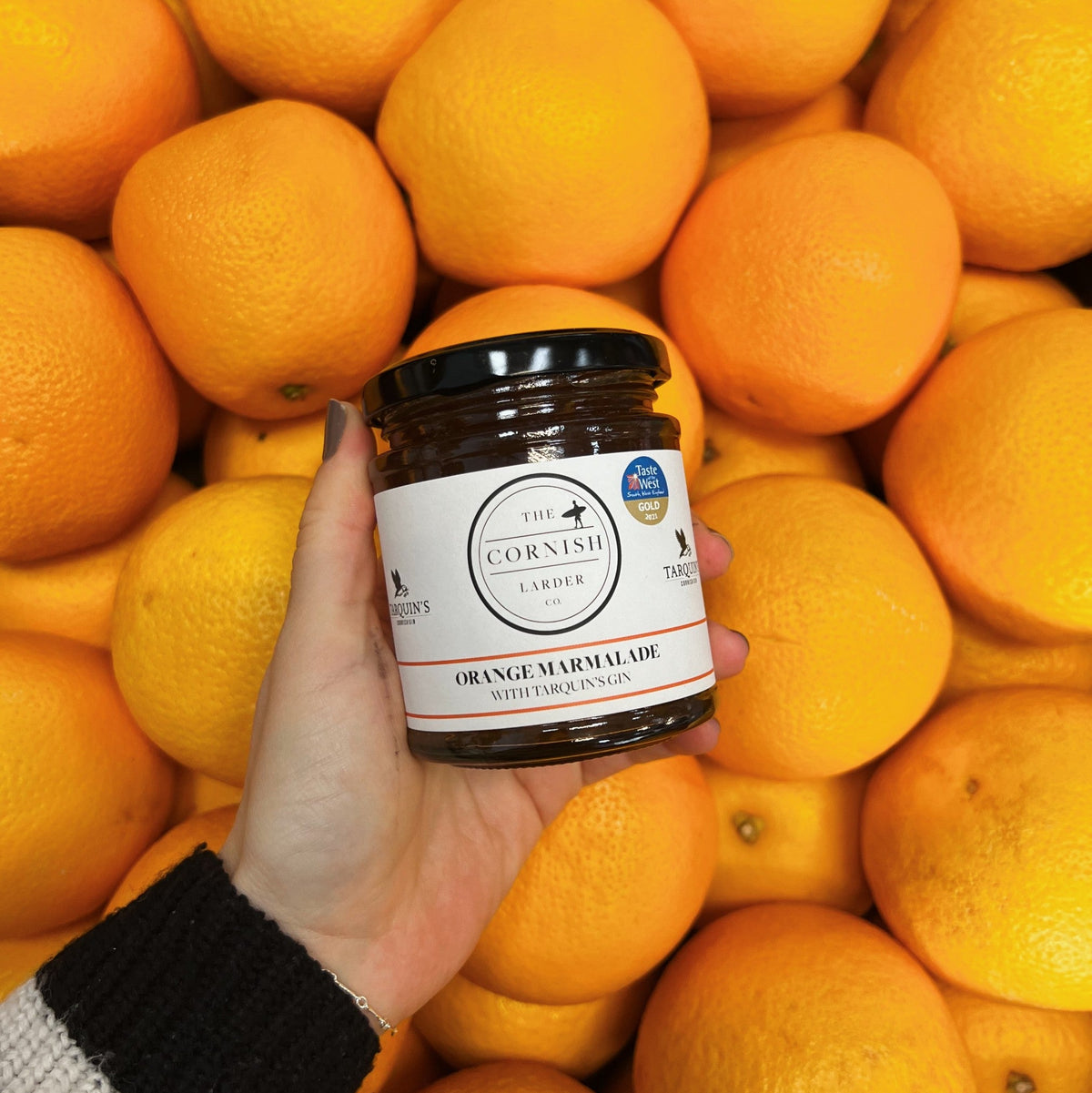 A hand wearing a black and cream knit sleeve holds a jar of The Cornish Larder Orange Marmalade in front of whole oranges.
